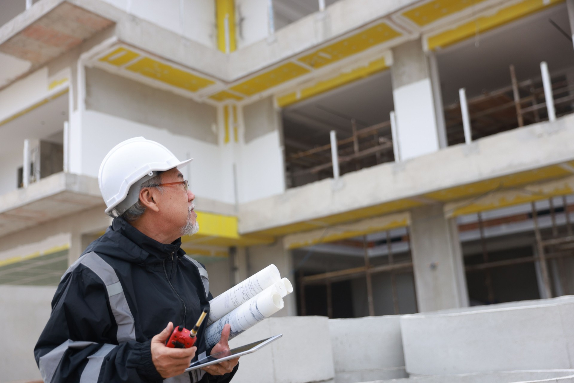 Senior engineer with black safety jacket holds the drawing and a digital tablet at the building construction site, infrastructure investment